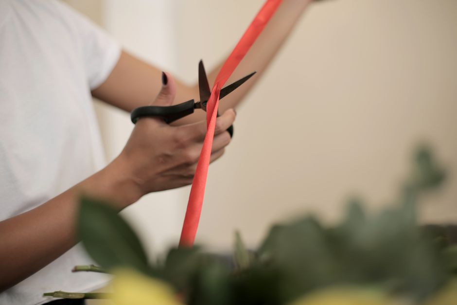 Close-up of a person holding black-handled scissors, carefully trimming a coral-colored ribbon around a bouquet of fresh flowers. The bouquet features a mix of vibrant yellow and green blooms, arrange