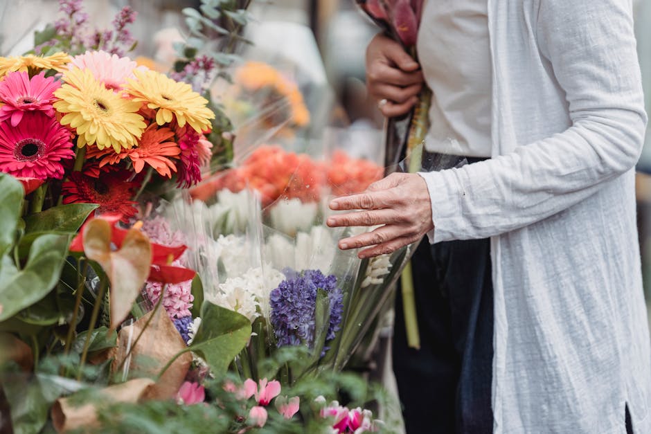 Close-up of a person dressed in a white jacket selecting vibrant, freshly cut flowers at a market stall. The bouquet features an assortment of colorful blooms including pink, yellow, orange gerbera da