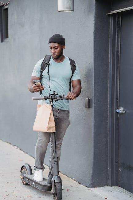 A young man with dark skin, wearing a black beanie, light blue t-shirt, and gray jeans, stands outdoors beside a gray wall, holding a smartphone in one hand and gripping the handlebars of an electric 