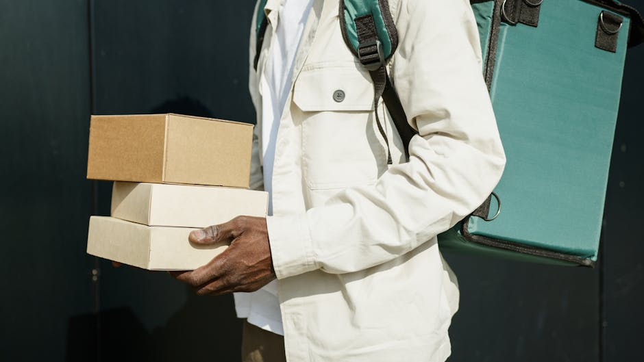 Close-up of a delivery person holding three boxes, with the top box being small and brown, and the other two boxes in shades of cream or white. The person is wearing a light-colored jacket and has a g