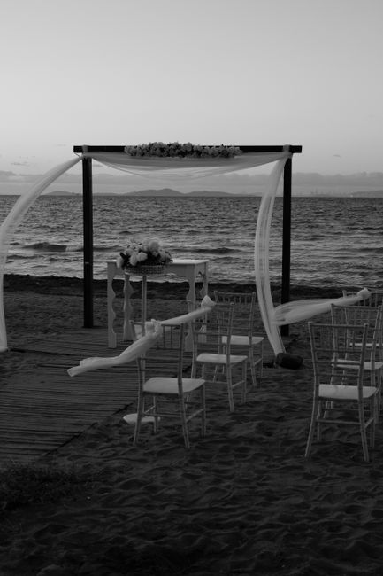 A seaside wedding setup featuring an outdoor floral arch draped with sheer white fabric, situated on a sandy beach with water and distant landforms visible in the background. In front of the arch, a s
