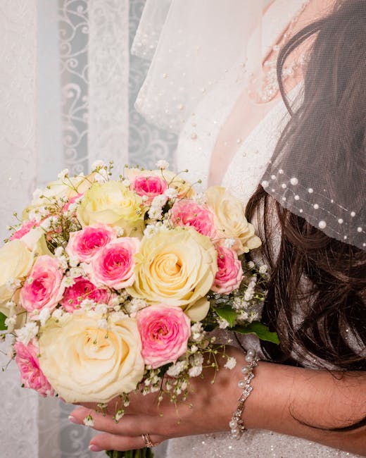 A bride's hand holding a bouquet arrangement featuring large cream-colored roses, pink spray roses, and delicate white baby's breath, complemented by green foliage. The bouquet is wrapped in light, el